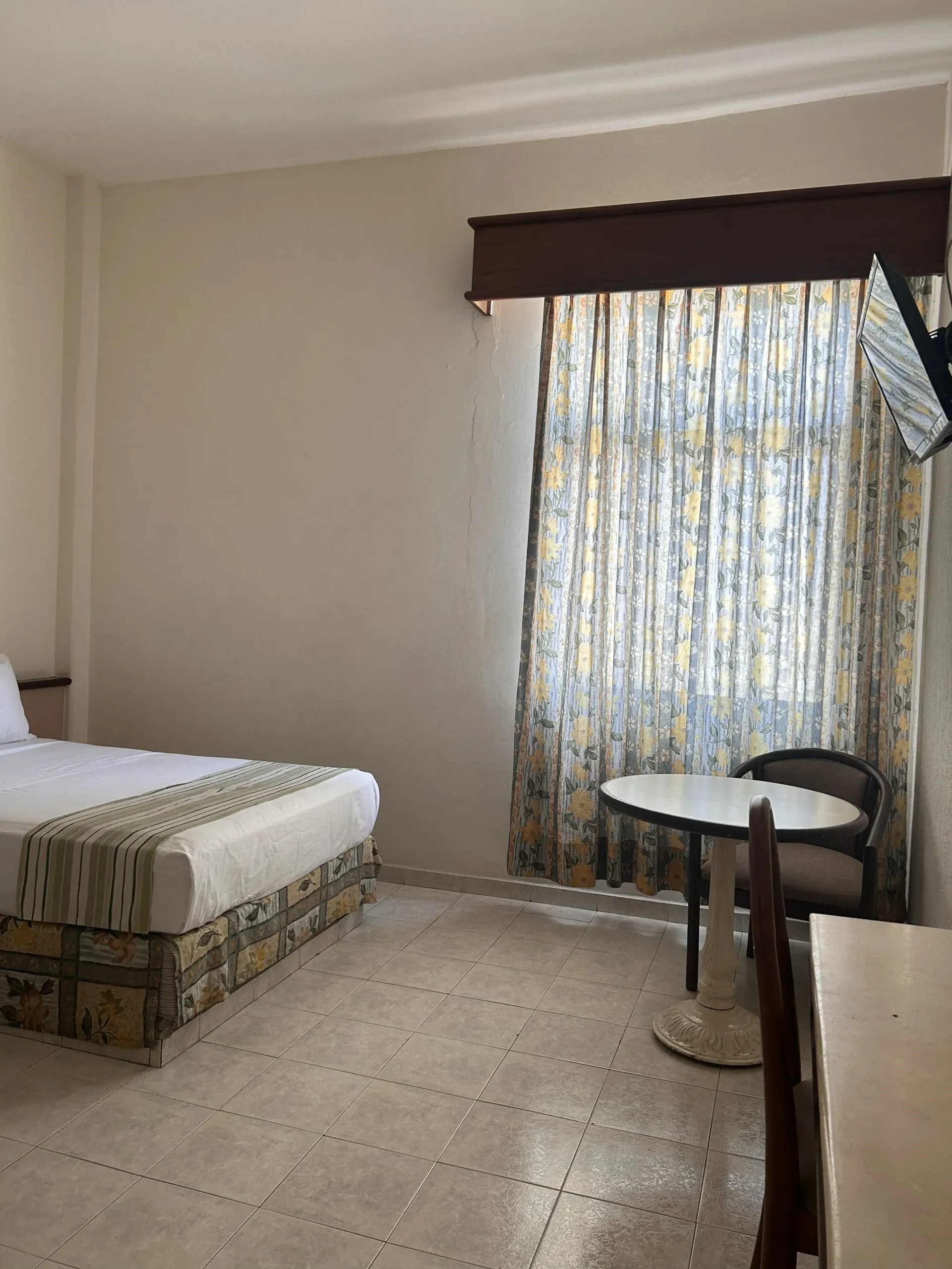 Una habitación de hotel con una cama bien hecha, una pequeña mesa redonda con una silla y una ventana con cortinas.