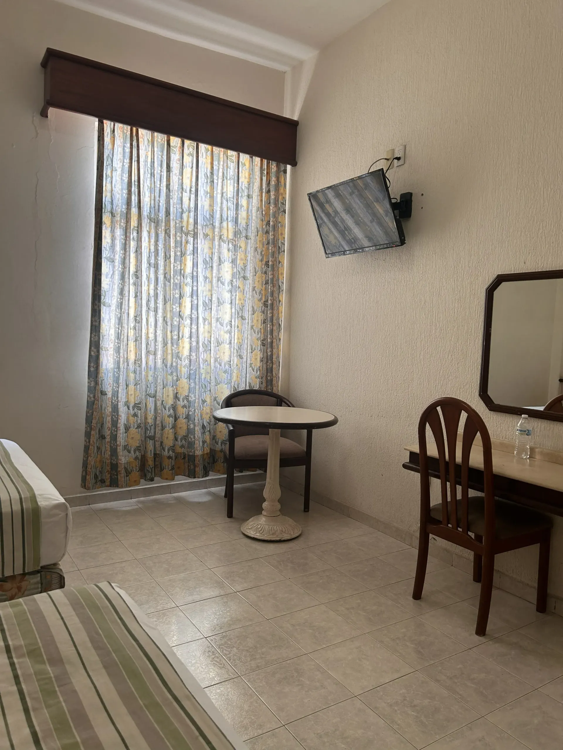 Una habitación de hotel con dos camas, una mesita redonda con una silla y un escritorio con espejo y televisión en la pared.