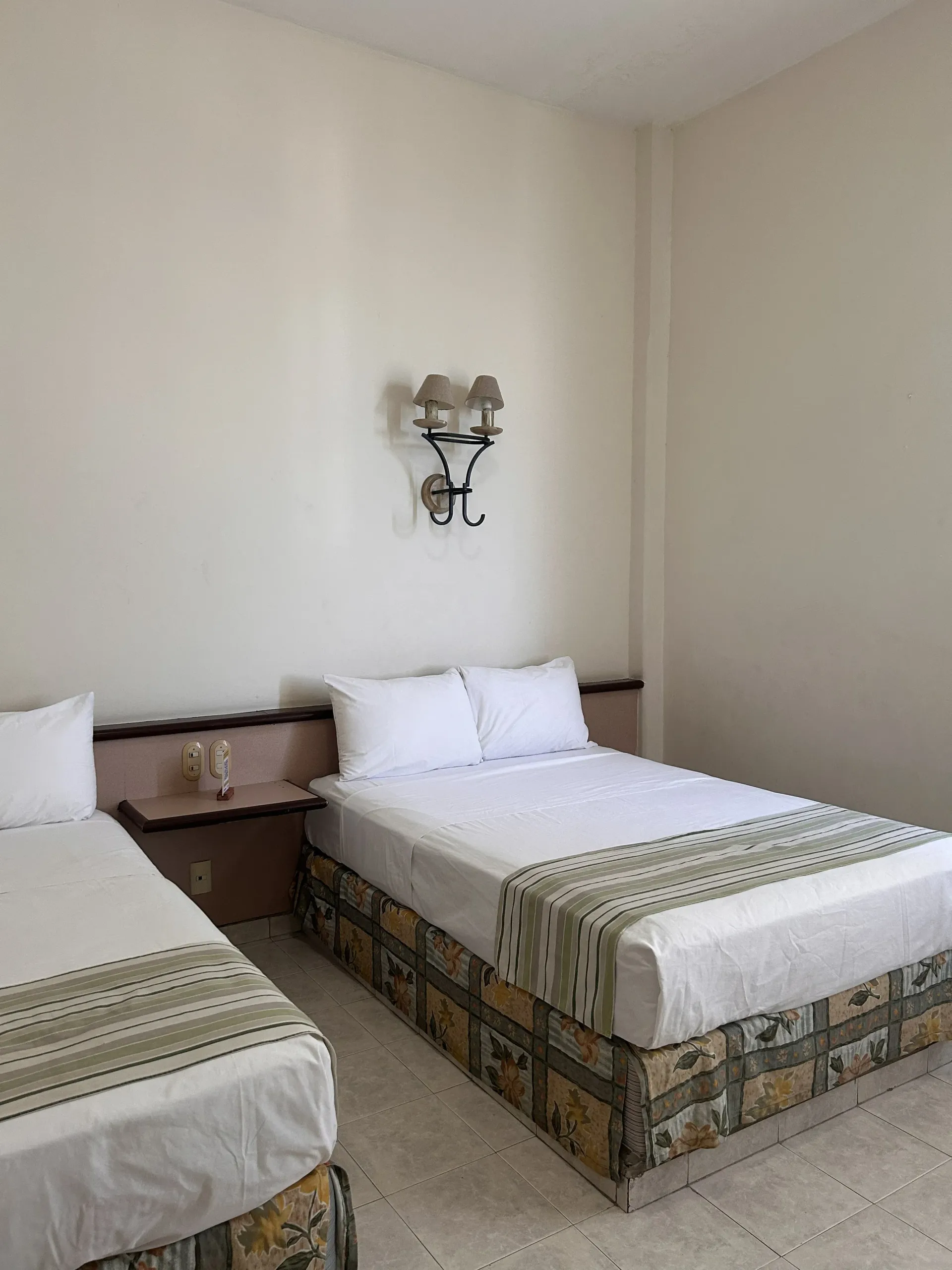 Una habitación de hotel con dos camas con sábanas blancas, colchas estampadas y una lámpara de pared sobre la cabecera.