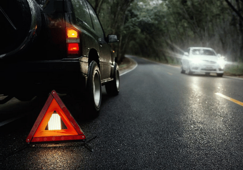 Car stopped on rainy road with hazard triangle; another car approaches with headlights on.