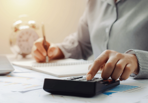 Person using a calculator while writing at a desk with papers and a clock in the background