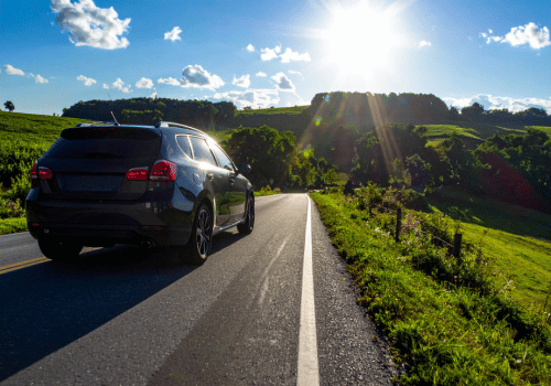 SUV driving along a sunny country road with green hills and bright sunlight in the background.