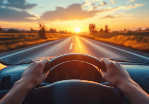 Hands on steering wheel driving toward a sunset on an open road