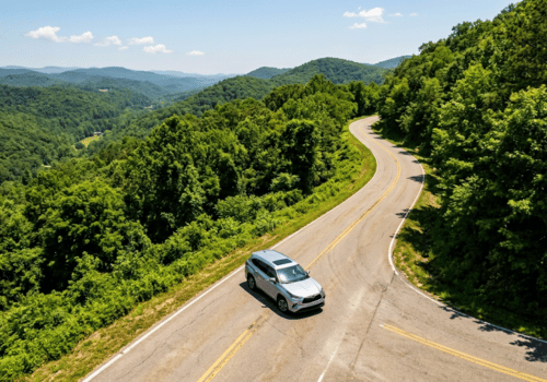 Silver SUV driving along a winding mountain road overlooking lush forested hills in summer