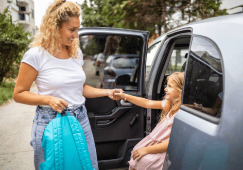 Woman handing a child’s bag through an open car door, with a blue skirt and pink dress visible.