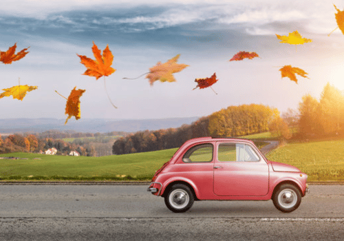 Red vintage car on a road with autumn leaves flying over a countryside landscape