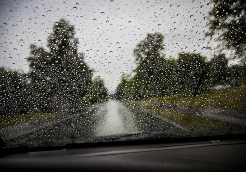Rainy road seen through a car windshield covered in raindrops, with trees lining both sides.