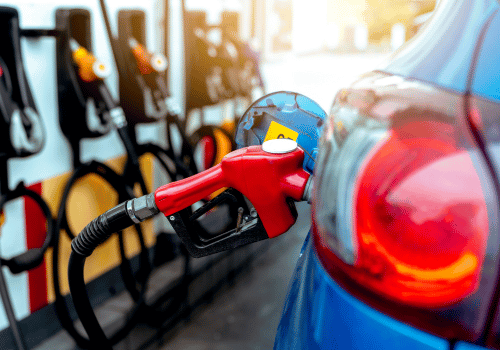 Red fuel pump nozzle inserted into a blue car at a gas station, with other pumps blurred in the background