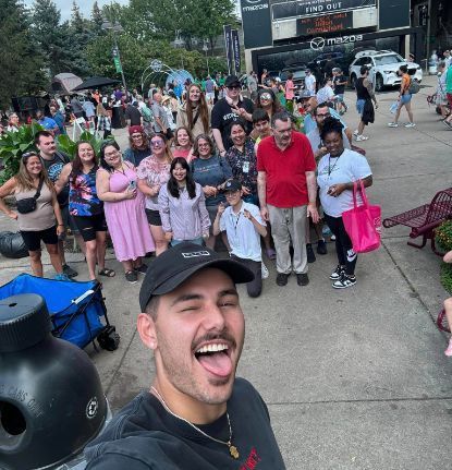 Man taking selfie with a large group of people outdoors, most smiling, near buildings and a parked wagon.