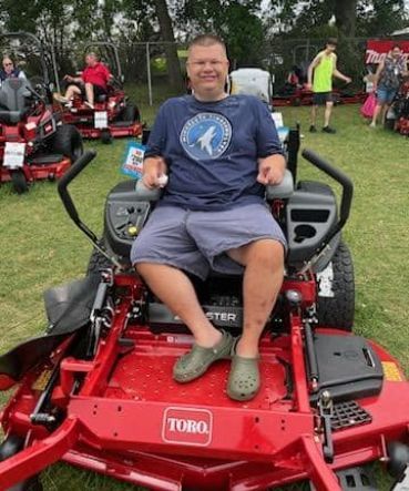 Man sitting on a red Toro riding lawnmower, smiling. Outdoors, surrounded by other mowers.