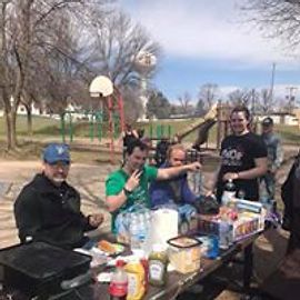 People at a picnic table with food and drinks in a park. Trees and a basketball hoop are visible.