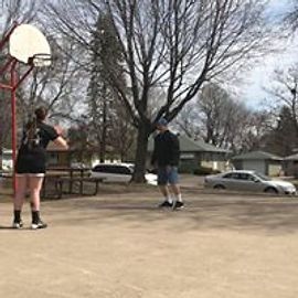 Two people playing basketball outdoors. One shooting, another standing near. Court, trees, cars in background.