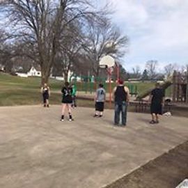 People playing basketball on an outdoor court near a park.