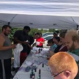 People tie-dyeing shirts under a white canopy. Various people are working at a table, applying dye.