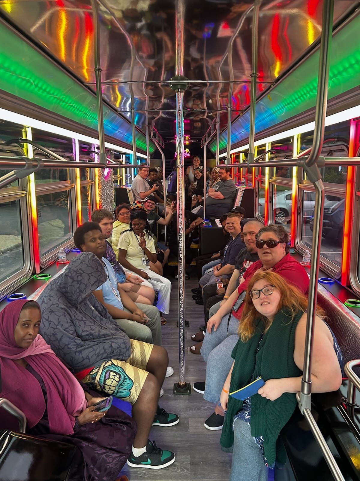 People sit inside a party bus with colorful lights. Some passengers smile, looking at the camera.