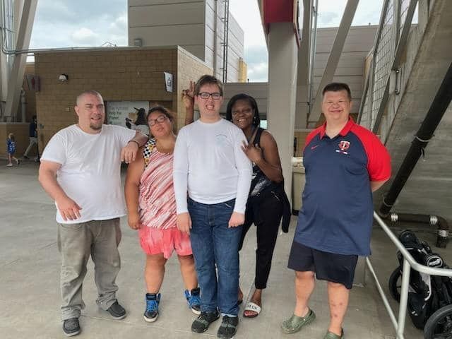 Group of people smiling outside a building. One gives a peace sign. They are under a partial roof structure.
