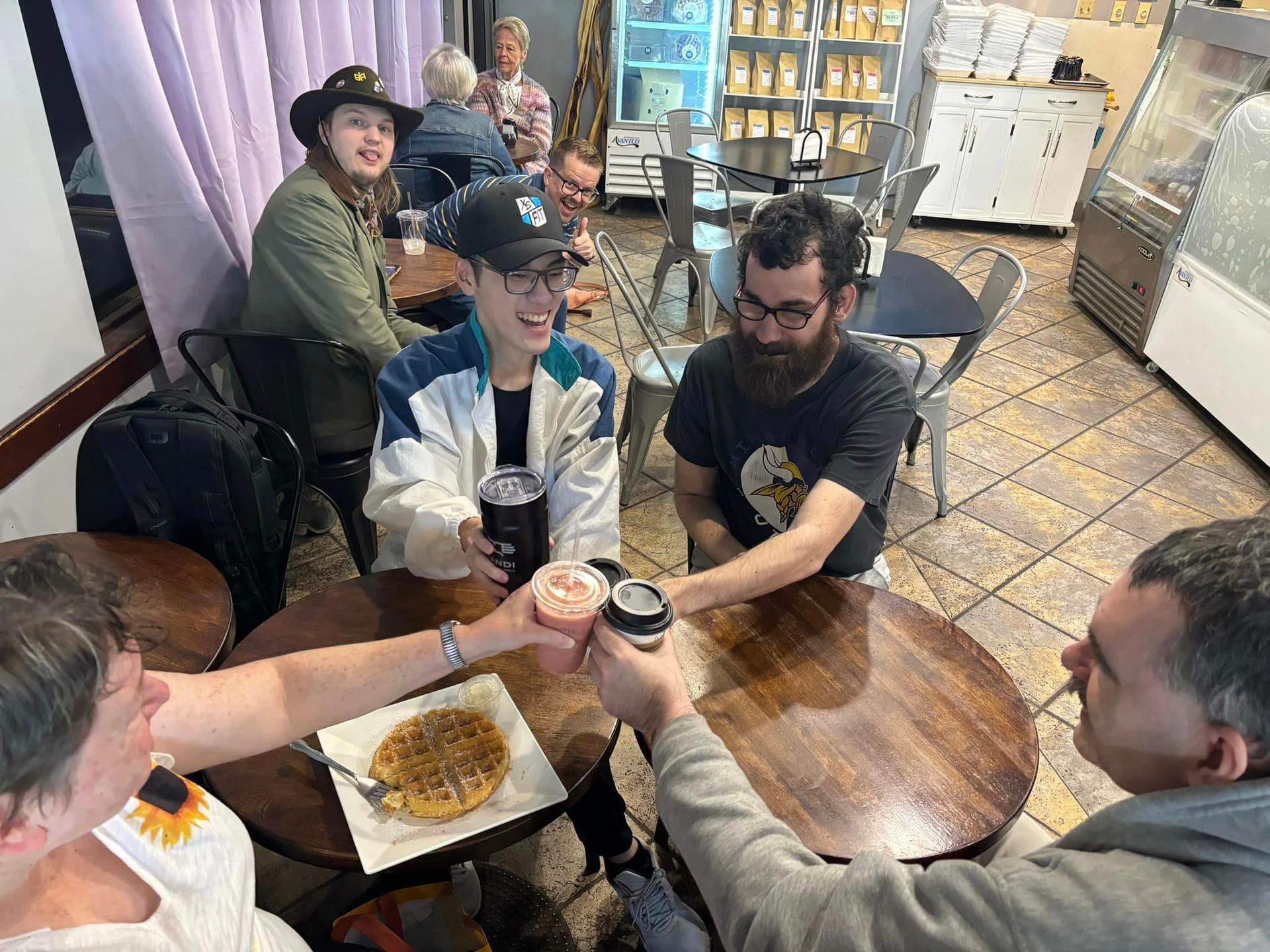 People at round tables in a cafe, toasting with drinks. One table has a waffle. Soft lighting.