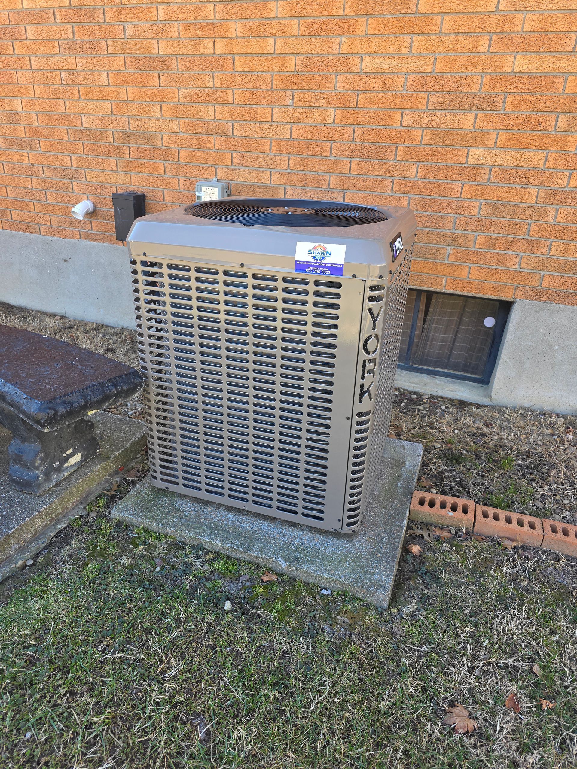 An air conditioning unit on a concrete base, set against a brick wall and a small window.