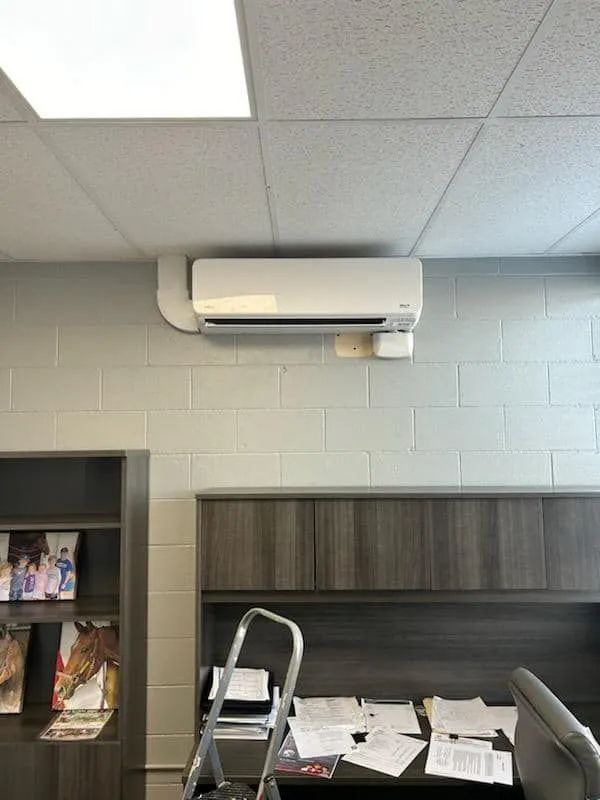 Office cubicle with papers scattered on desk, ladder, bookshelf, and wall-mounted air conditioner above.