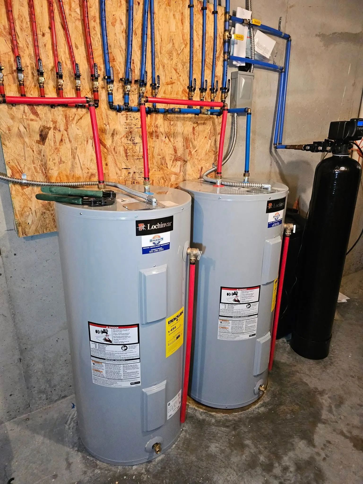 Two gray water heaters with colorful pipes in a utility room, next to a black tank.