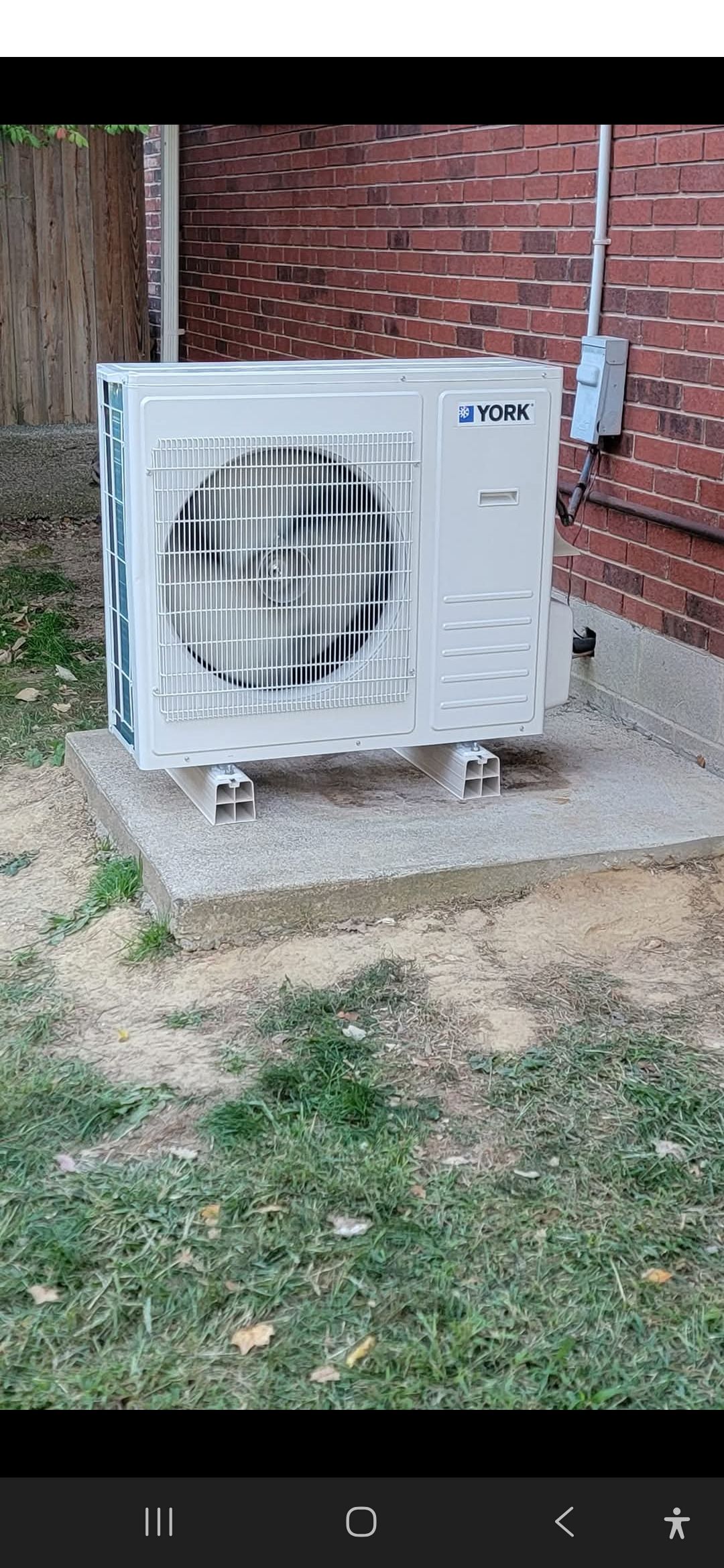 A white air conditioning unit on a concrete pad near a brick wall, situated in a yard with grass and mulch.
