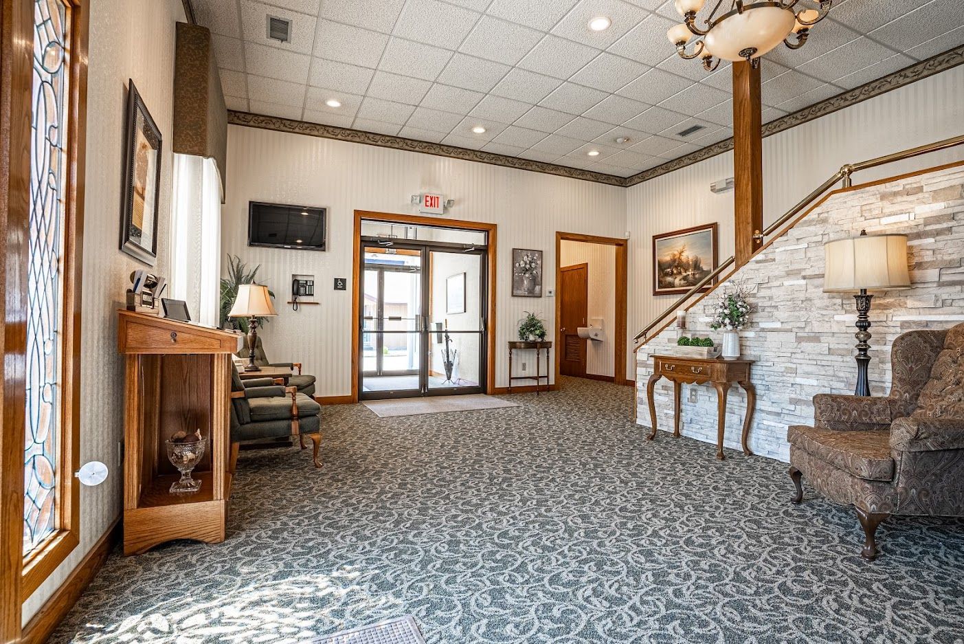 Lobby with patterned carpet, wood trim, staircase, chairs, and glass entry doors under a chandelier.