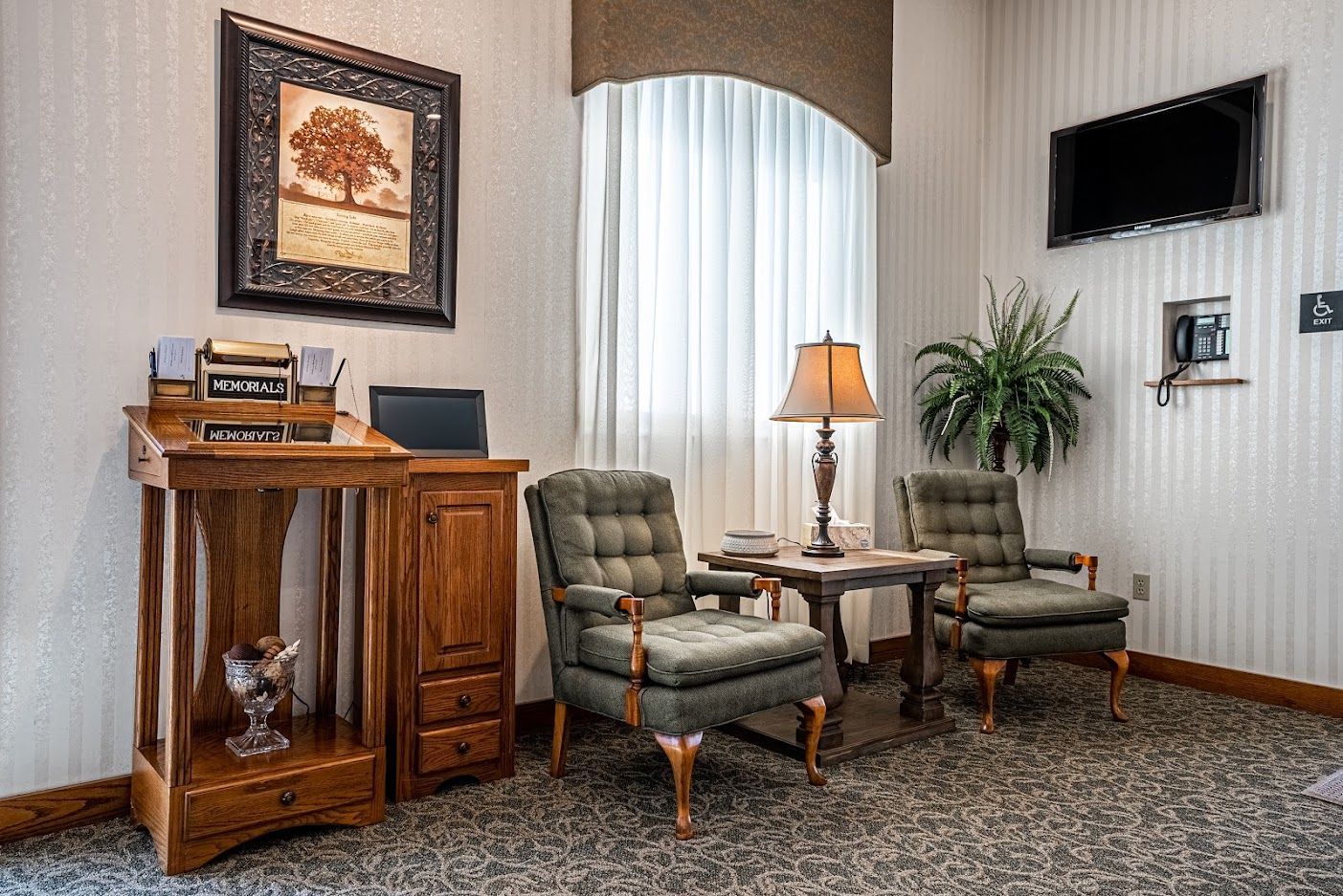 Quiet lobby seating area with two green armchairs, small table, lamp, cabinet, and wall-mounted TV