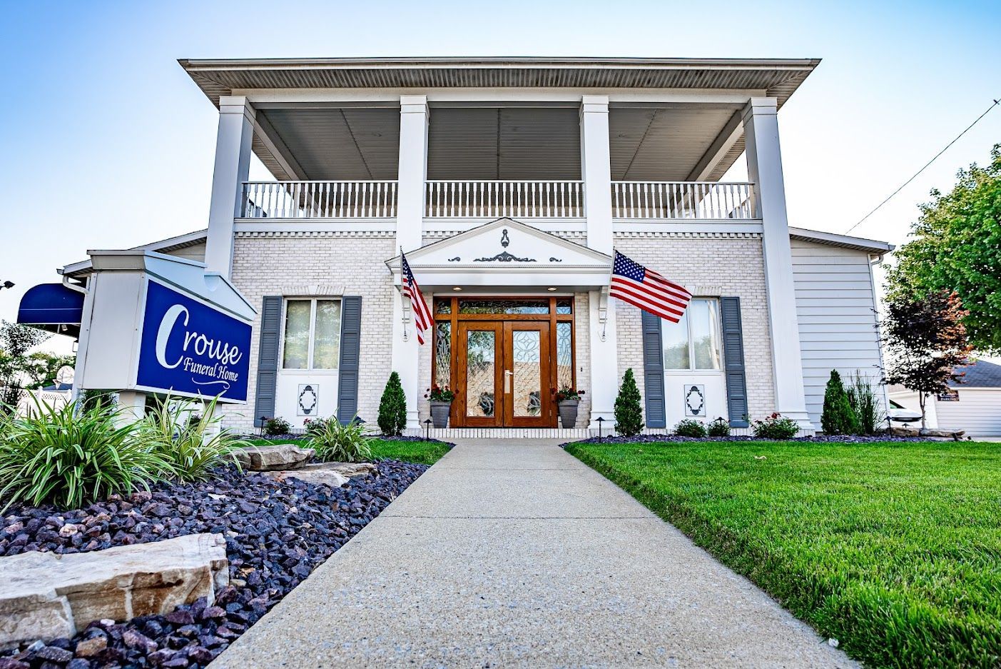 White building with columns, American flags, and a blue sign beside a landscaped walkway