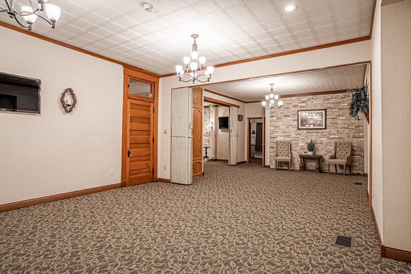 Empty carpeted lobby with wood doors, chandeliers, wall art, and seating area ahead