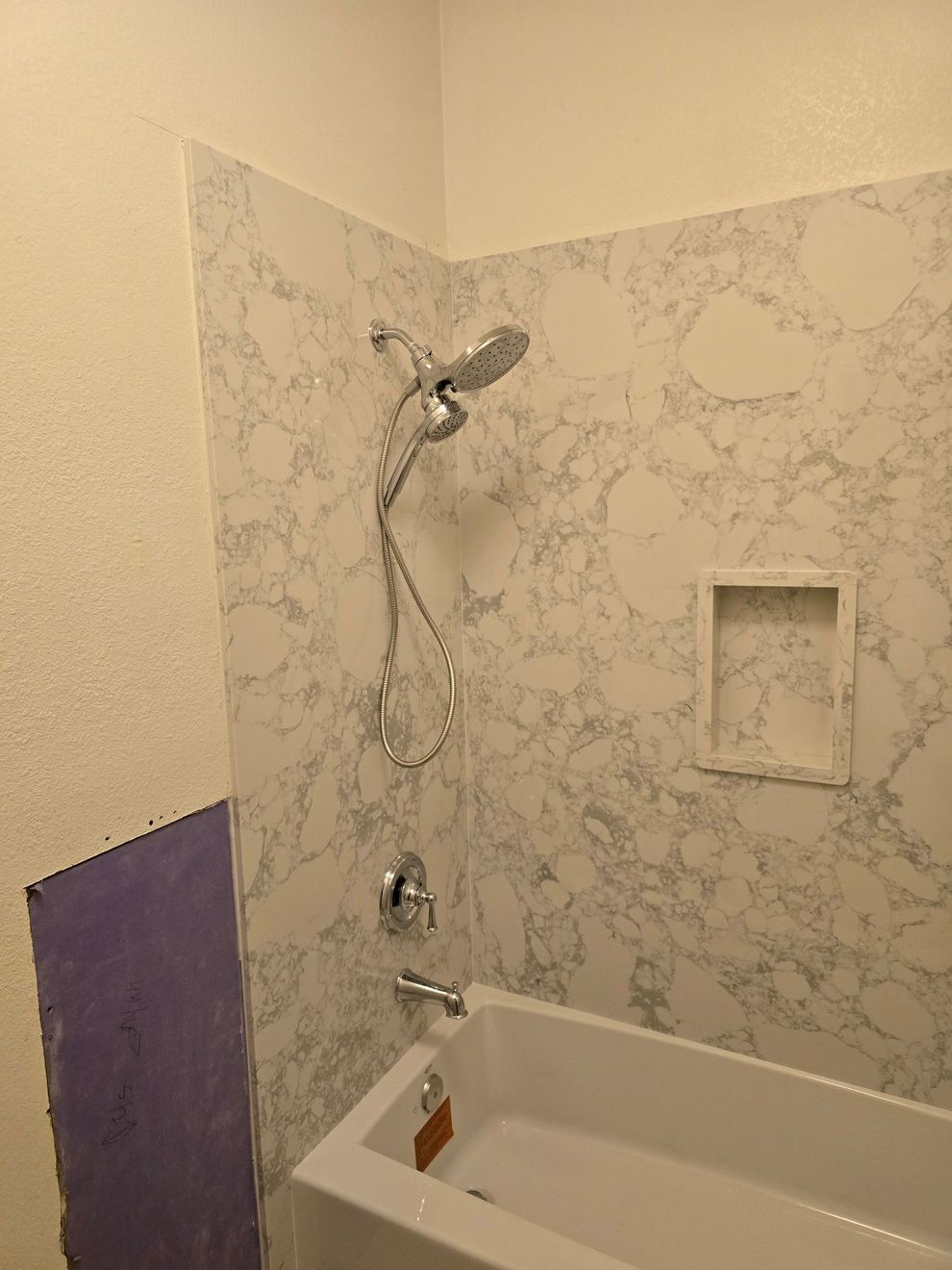 Bathroom shower over bathtub with marble-patterned wall and built-in shelf niche