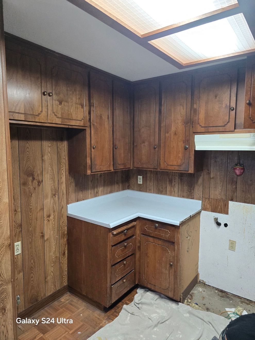 Small wood-paneled kitchen corner with brown cabinets, white countertop, and fluorescent ceiling light.