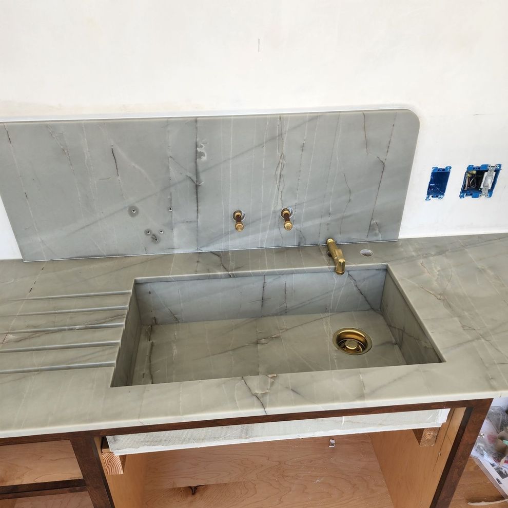 Marble kitchen sink and countertop with brass faucet under construction, against a white wall.