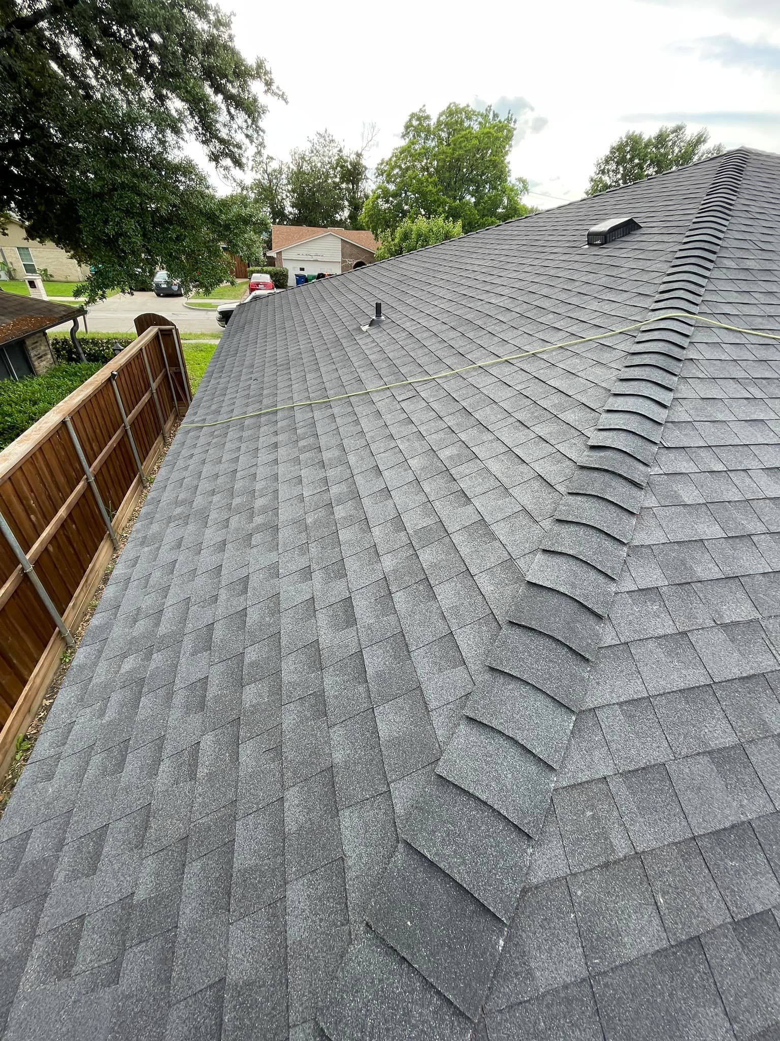 Dark gray asphalt shingle roof of a house. A wooden fence is on the left side, trees in the background.