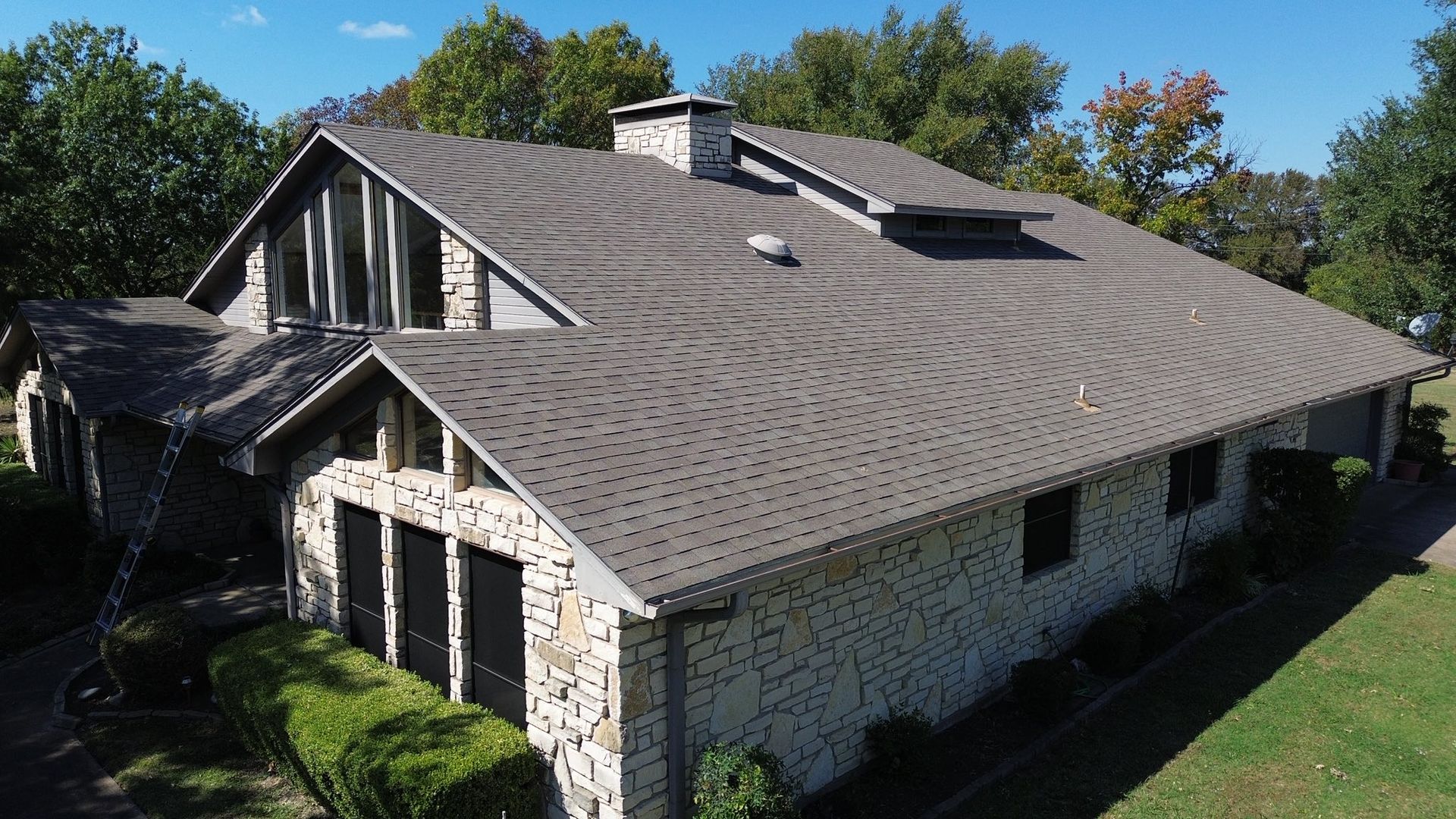 Aerial view of a suburban house with a gray shingle roof, brick walls, and surrounding trees.