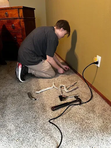 Person kneeling on carpet near wall outlet, adjusting tangled electrical cords beside a desk.