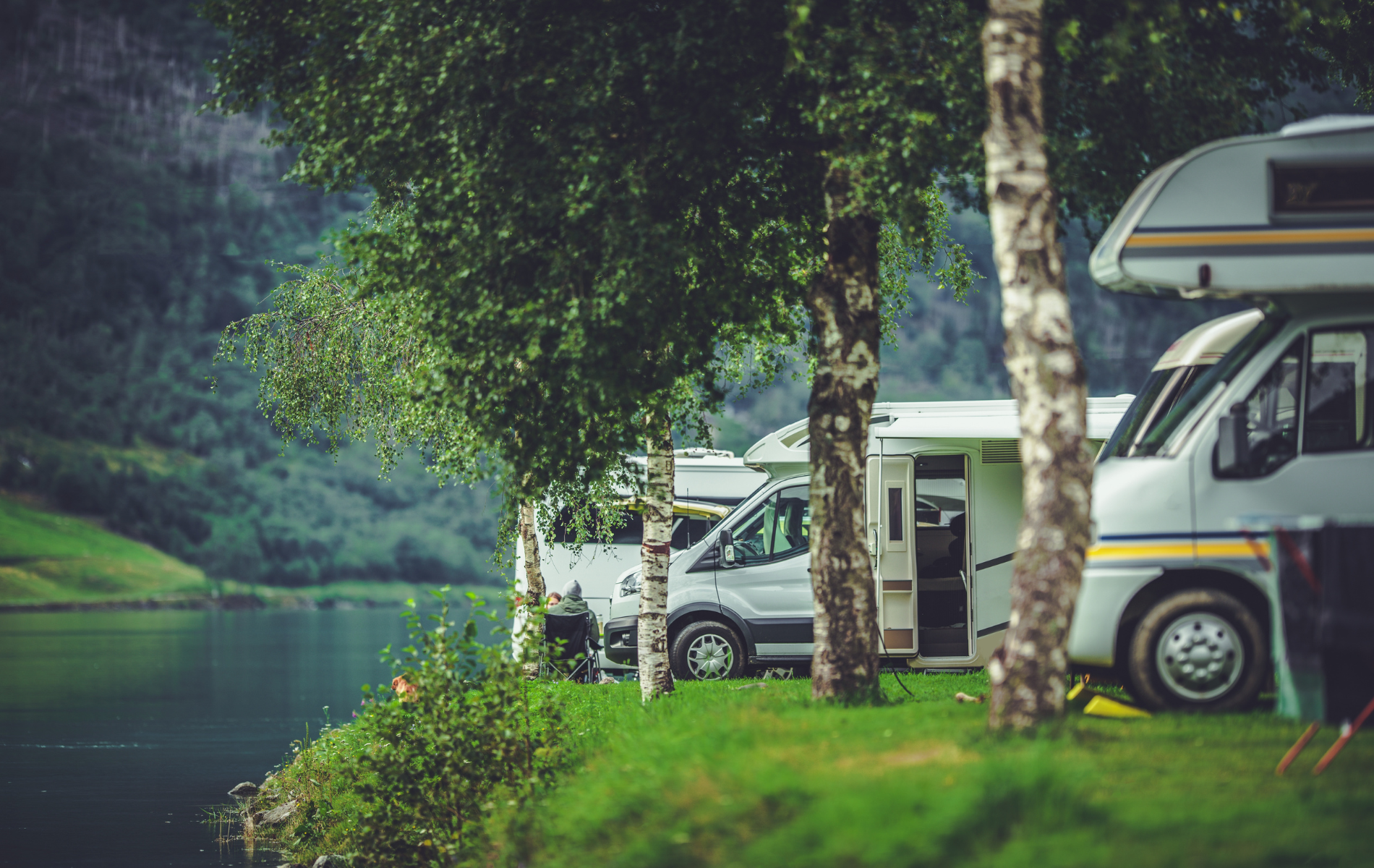 Campers parked near a lake and trees; green grass and mountains in the background.