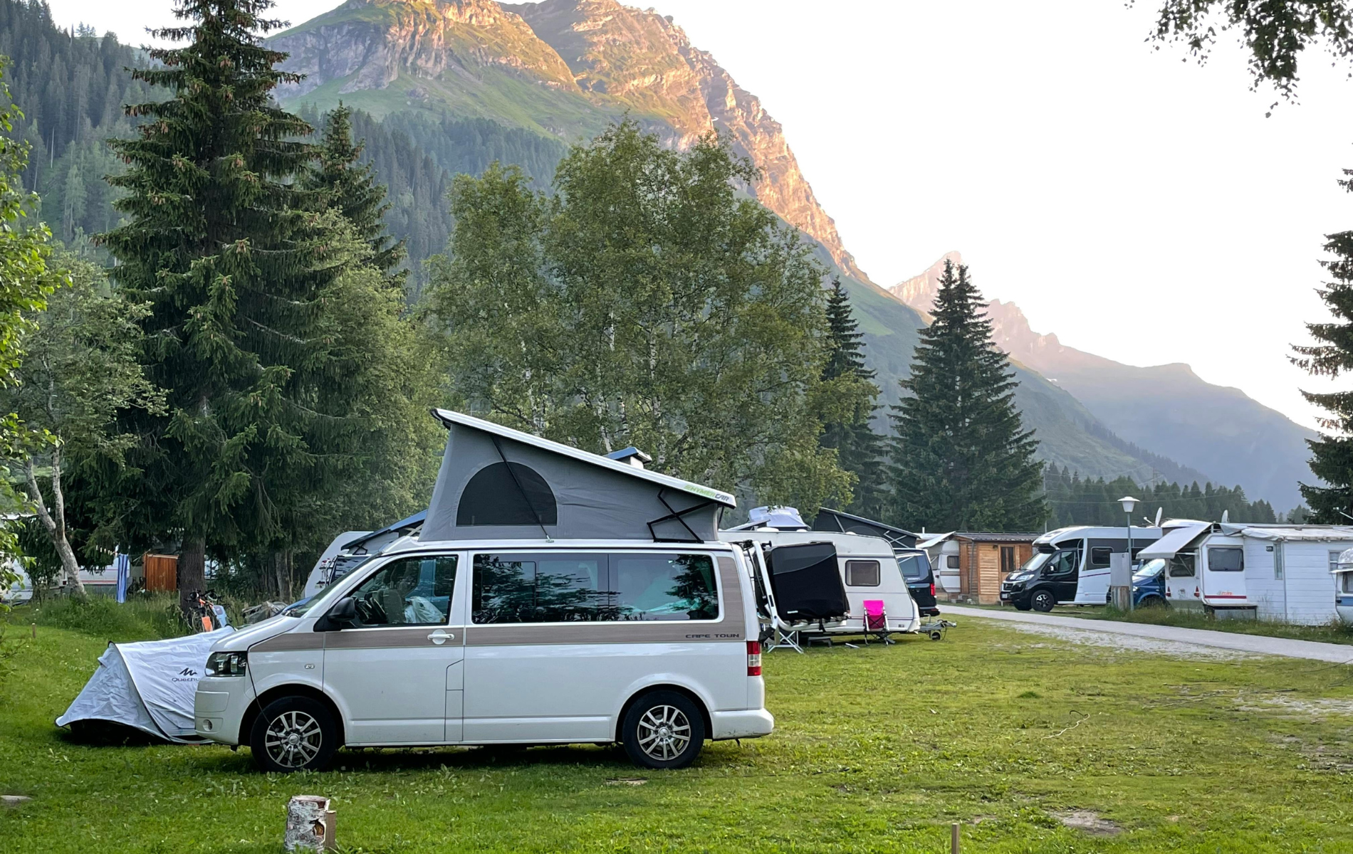 Camping ground with camper vans and tents, set against a backdrop of mountains and trees.