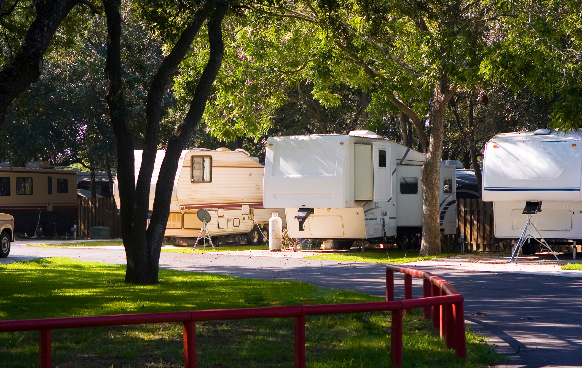 Campground with RVs parked under trees, with green grass and a red fence.