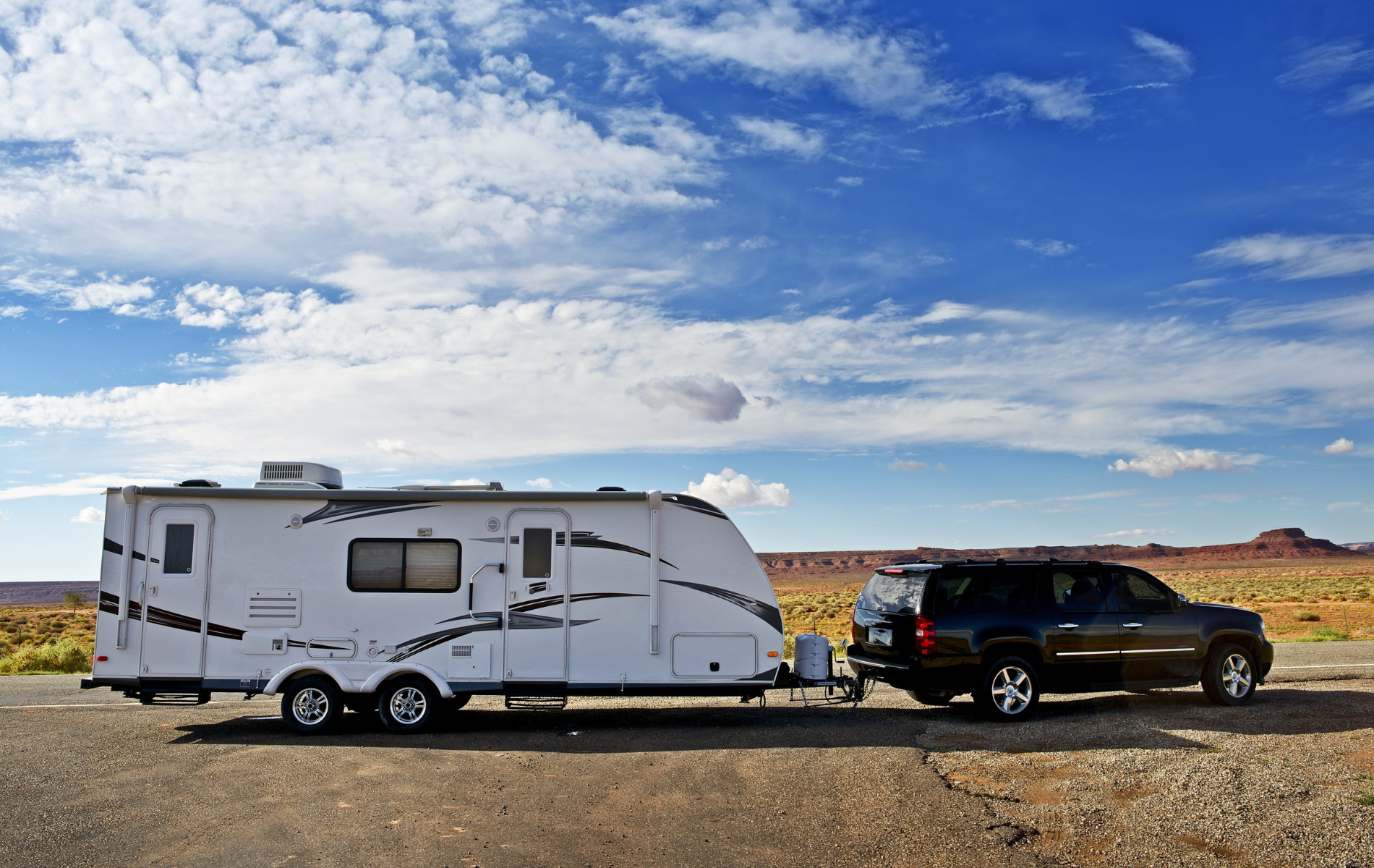 Black SUV towing a white travel trailer on a desert road under a blue sky with clouds.