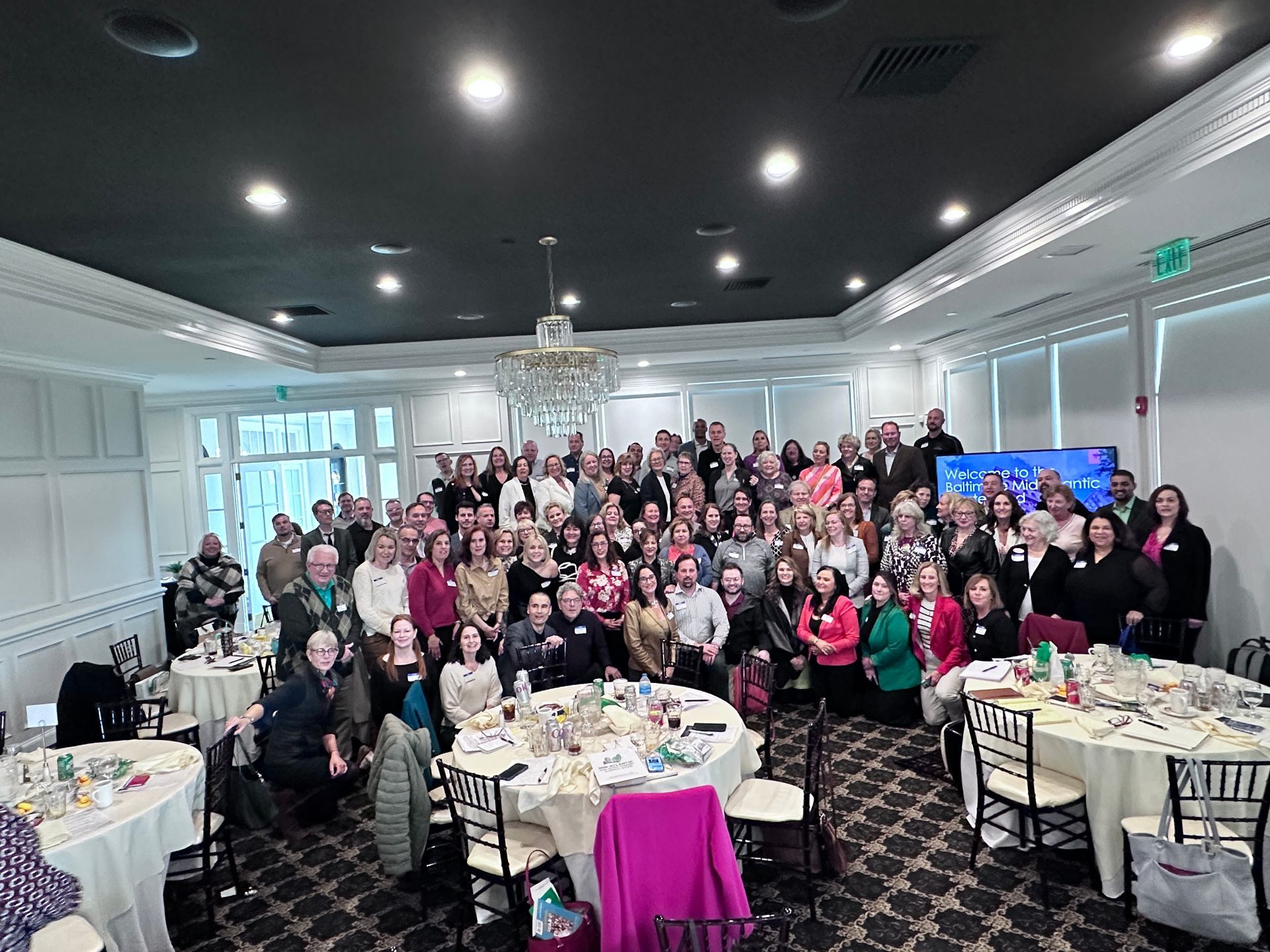 Large group posing in a bright banquet hall with round tables and a chandelier, many smiling and waving