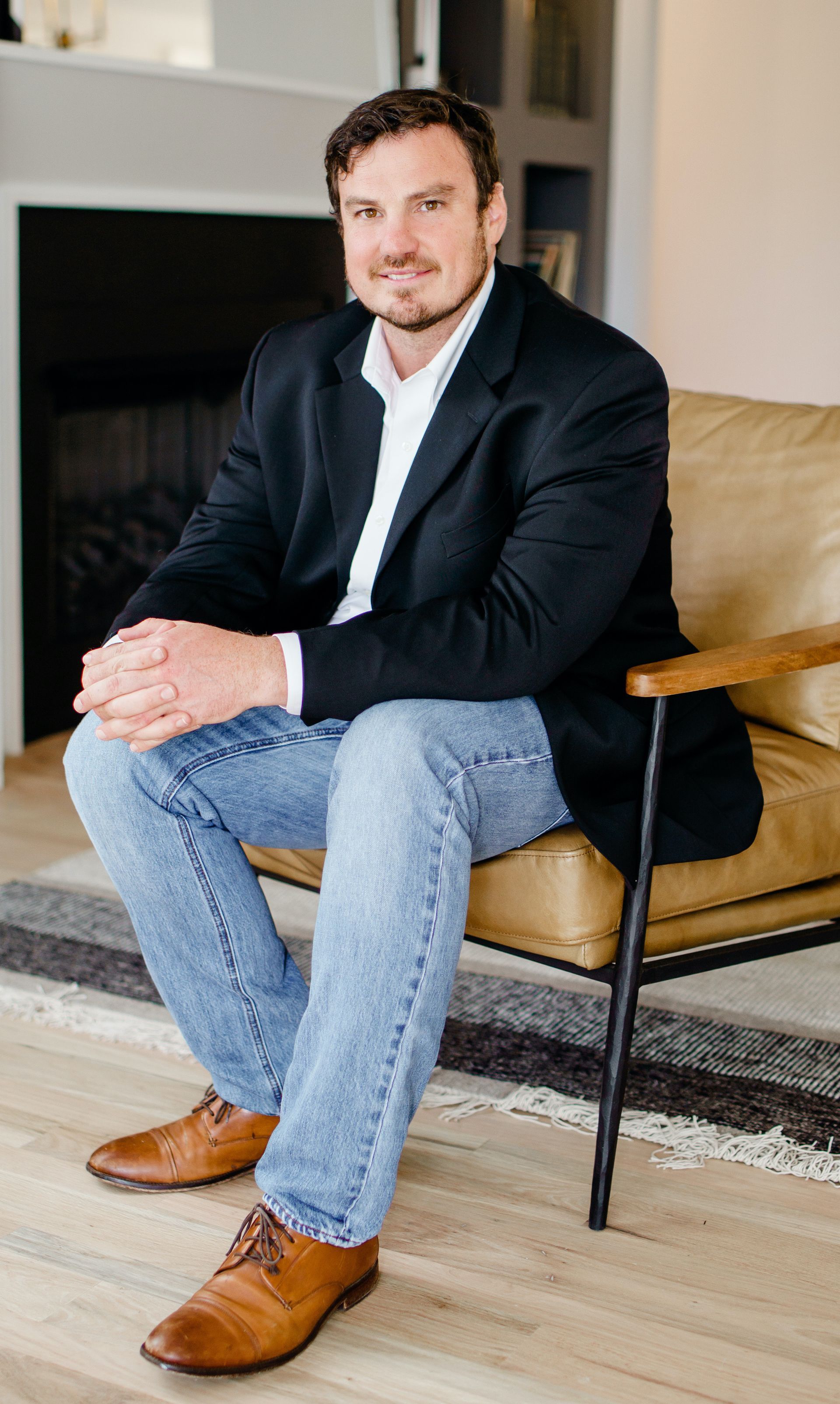Man seated on a chair indoors, wearing a dark blazer, jeans, and brown shoes, with hands clasped.