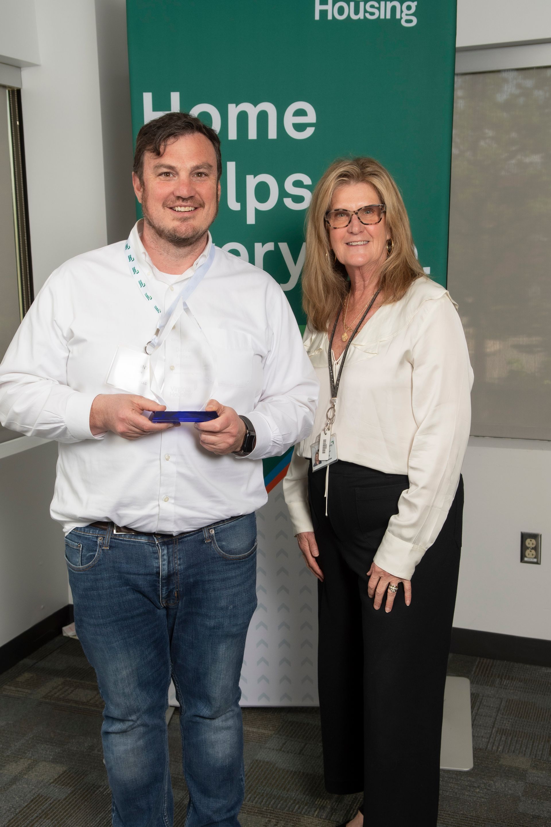 Two people standing indoors in front of a green “Home” banner, holding a small award plaque.