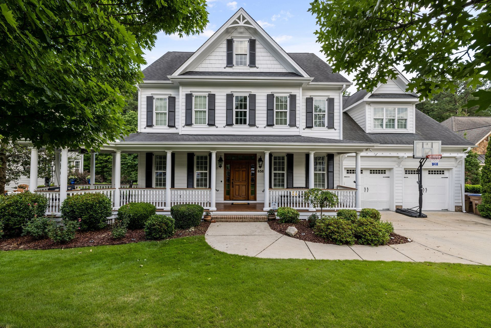 Gray two-story house with white trim, front porch, manicured lawn, and driveway under leafy trees