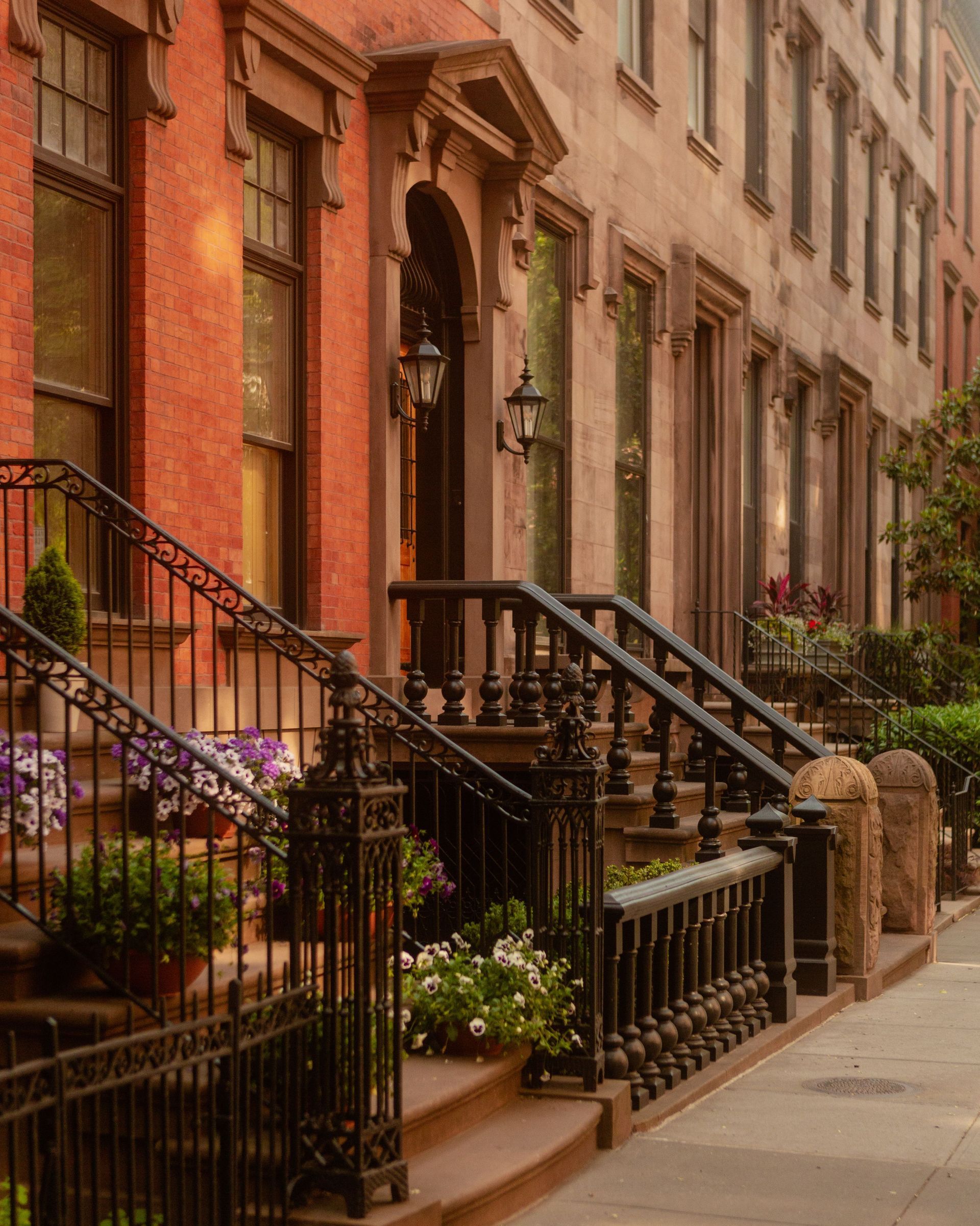 Brownstone rowhouses with black iron stoops and flowered railings along a city sidewalk
