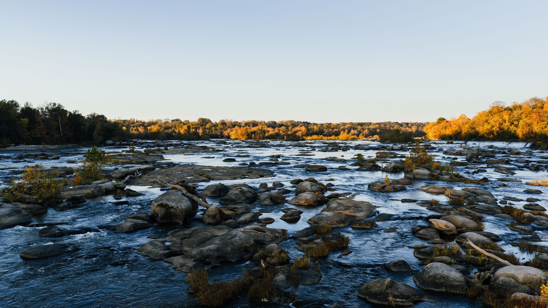 Snow-dusted rocky riverbed at sunset with dark trees and golden hills in the distance