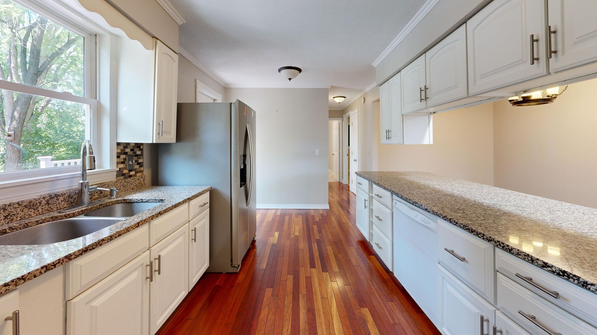 Bright galley kitchen with white cabinets, granite counters, stainless fridge, and hardwood floors.