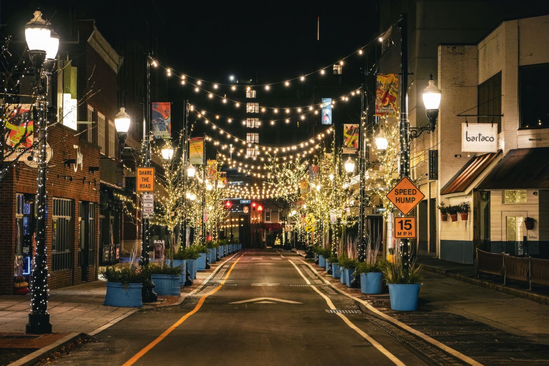 Empty, lamp-lit street at night with string lights, blue planters, and buildings lining both sides