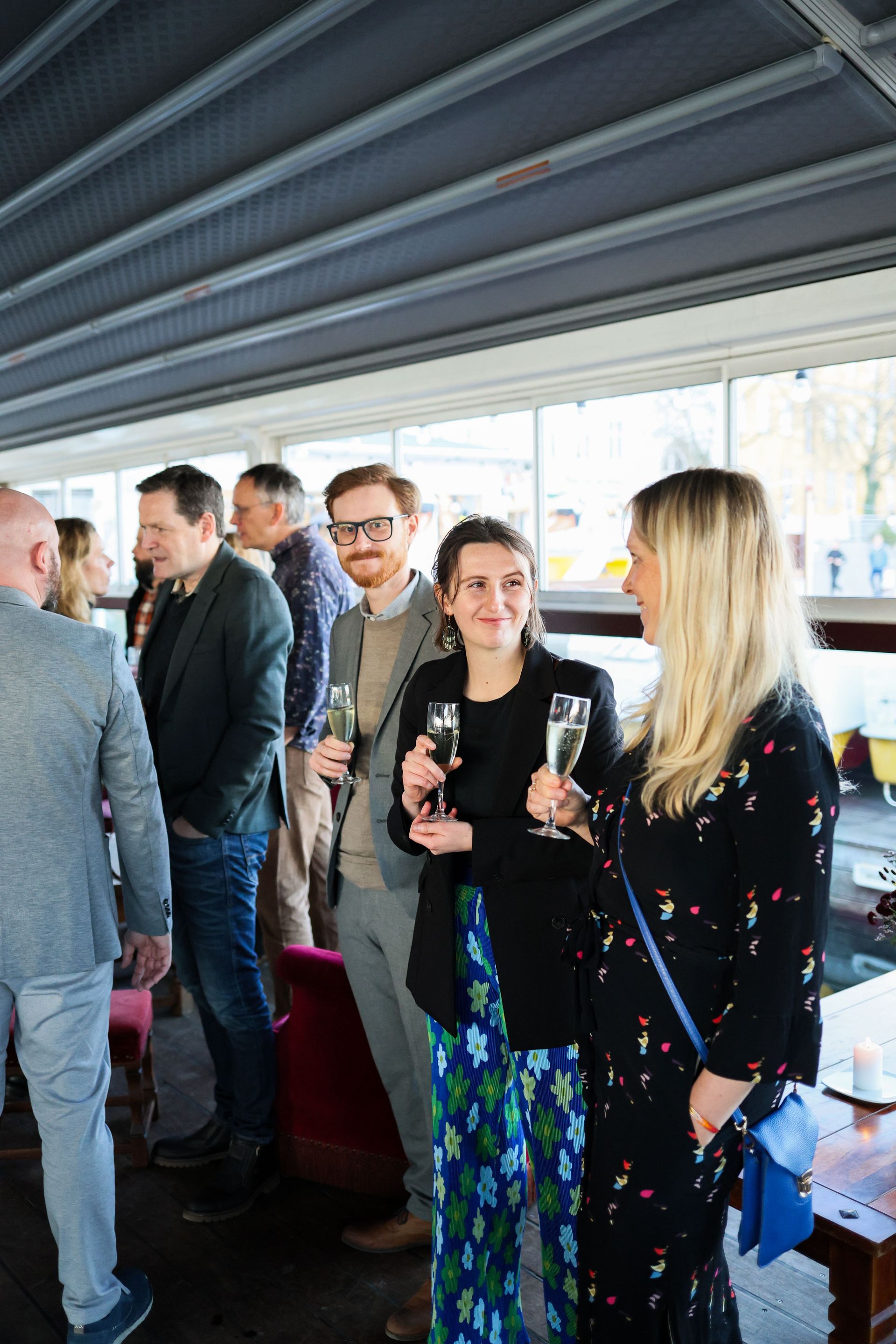 People chatting at an indoor event, holding drinks near large windows and a metal ceiling.