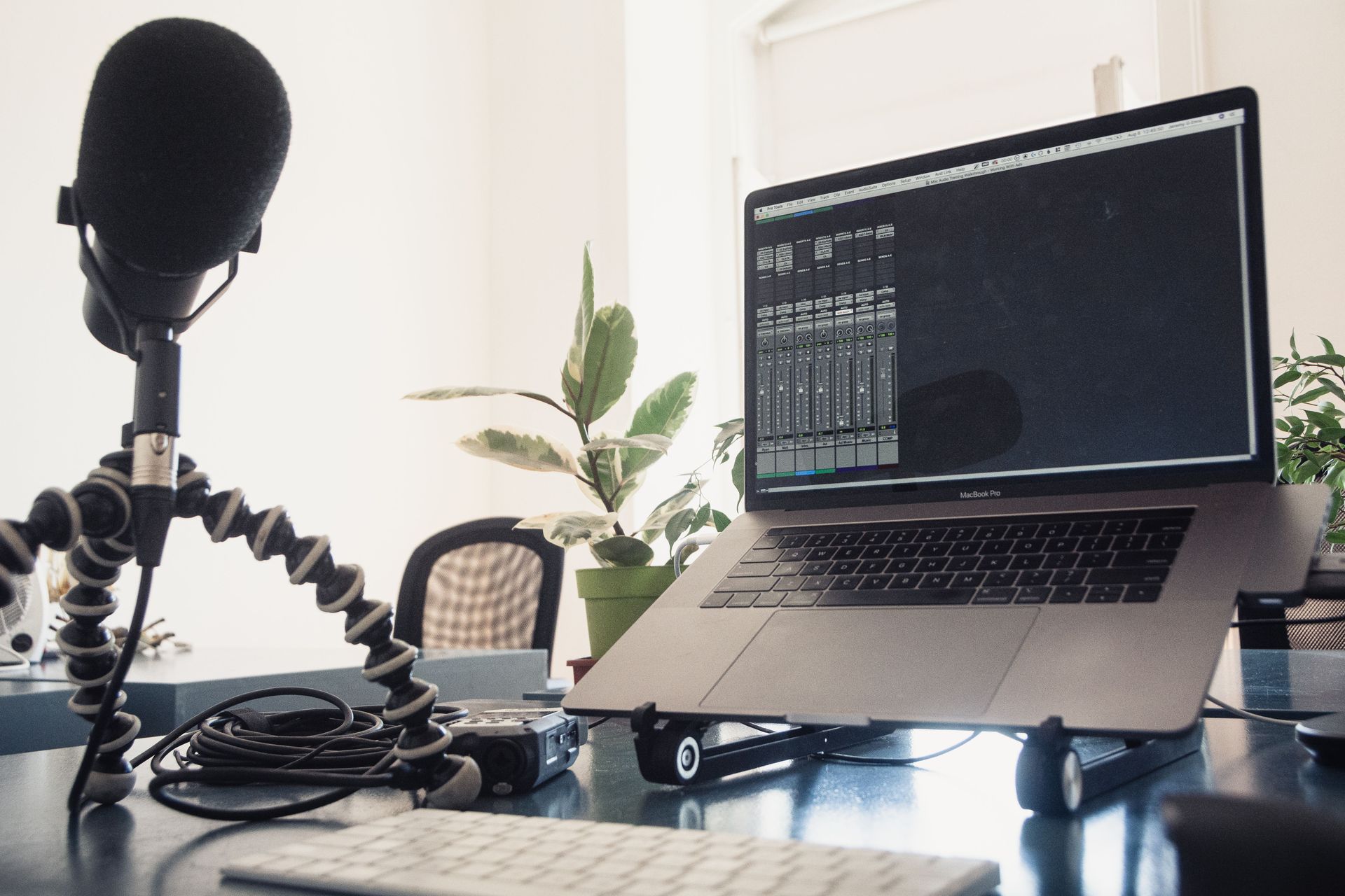 Home podcast setup with laptop, microphone, and tripod on a desk by a plant.
