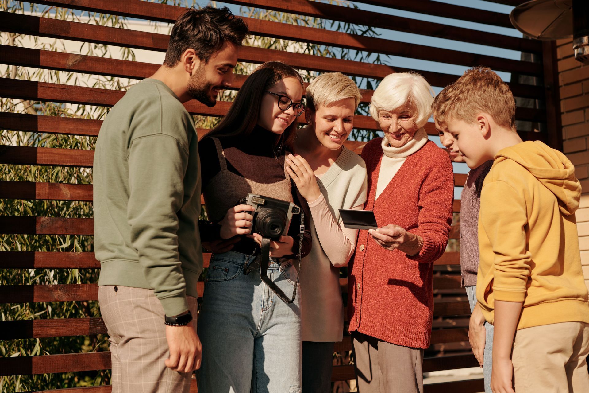 Five people smiling together outdoors by a wooden slat wall, looking at a phone or camera.